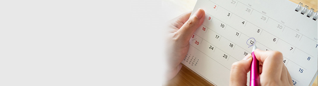 Mulher de óculos, vestindo uma camisa azul escura, se inclina sobre uma cama branca com um sorriso, parecendo pensativa. / Close de um calendário com a mão de uma mulher marcando um dia com uma caneta.
